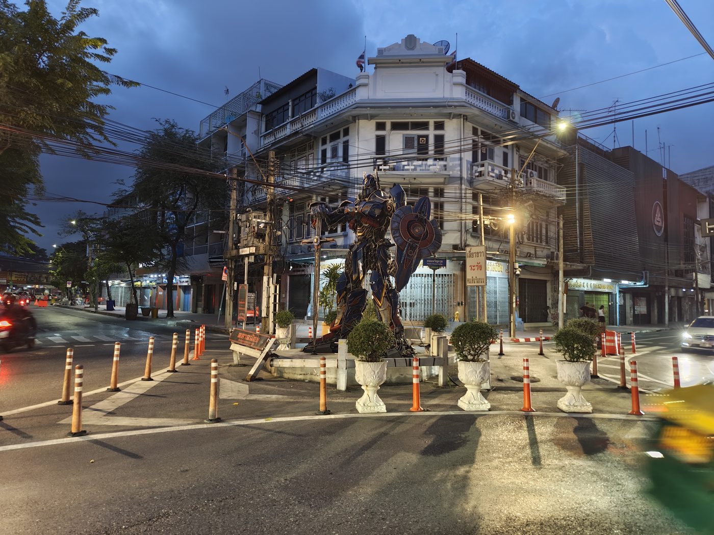 A scrap-metal Transformer on a traffic island in front of an Art Deco corner shophouse, at dusk. Bangkok does not need permission to be interesting.